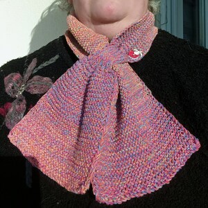 Peut inclure: Une écharpe tricotée multicolore avec un schéma de couleurs rose, bleu et jaune. L'écharpe est nouée en un nœud lâche autour du cou.
