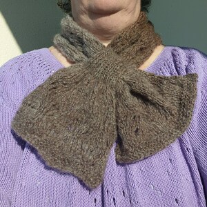 Peut inclure: Une écharpe en tricot marron avec un nœud noué à l'avant. L'écharpe est portée par une personne portant un pull violet.