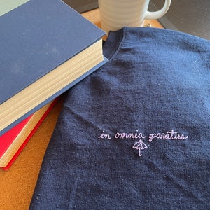 Op de afbeelding: Een marineblauw sweatshirt met de geborduurde zin "in omnia paratus" en een paraplu-ontwerp. Een witte mok met een zwarte rand en twee hardcover boeken staan ook in de afbeelding.