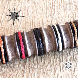 Könnte beinhalten: Nahaufnahme eines braunen Lederarmbands mit fünf verschiedenen farbigen Lederriemen. Die Riemen sind orange, rot, schwarz, weiß und braun. Das Armband ist aus echtem Leder und trägt ein "Vera Pelle"-Etikett.