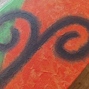 May include: A close-up of a handmade journal with a textured orange and green cover. The cover features a black painted swirl design.