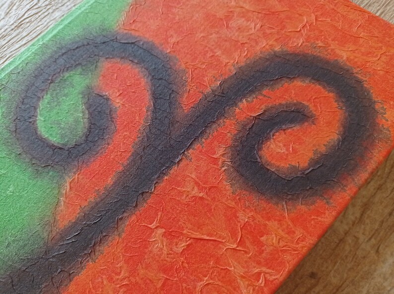 May include: A close-up of a handmade journal with a textured orange and green cover. The cover features a black painted swirl design.