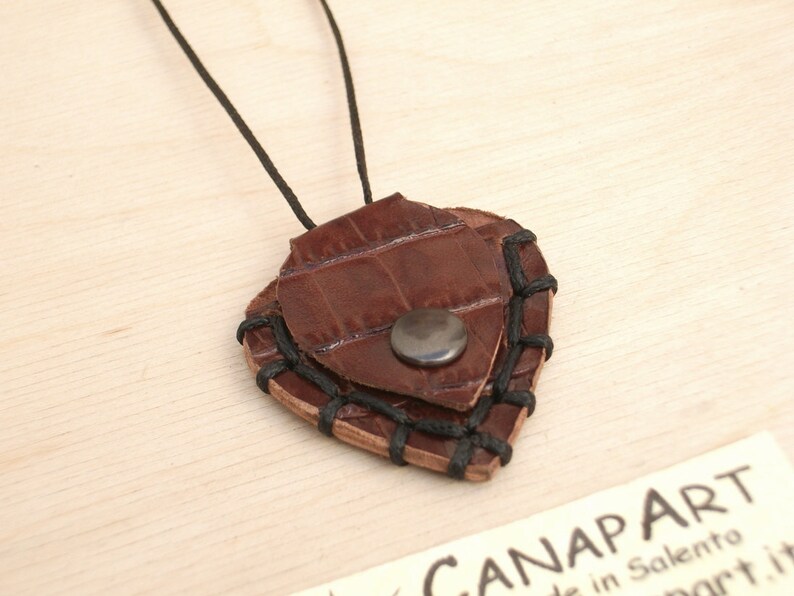 May include: A brown leather guitar pick necklace with black stitching and a silver button. The pick is shaped like a heart.