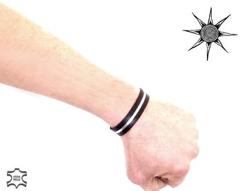May include: A black leather bracelet with a white stripe is worn on a wrist. The bracelet is made of genuine leather, as indicated by the "Vera Pelle" stamp. A black and white sun graphic is in the background.