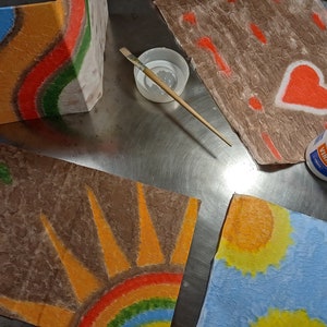 May include: Four pieces of paper with different designs painted on them. The designs include a rainbow, a sun, a heart, and a series of lines. The paper is brown and textured. There is a bottle of glue and a paintbrush on the table.