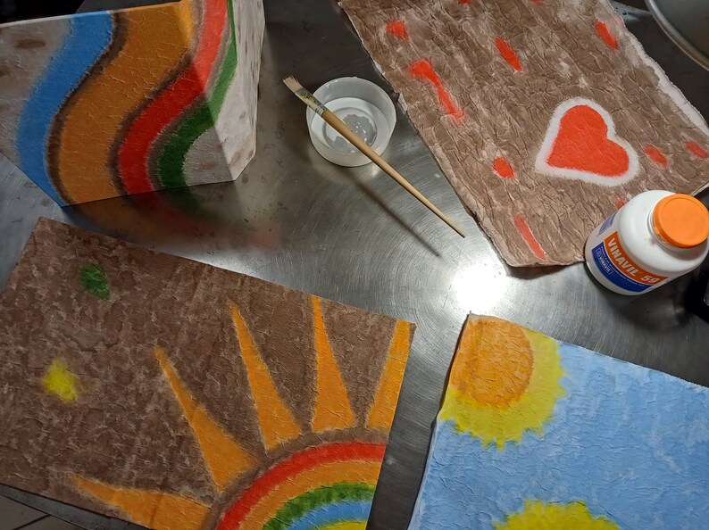 May include: Four pieces of paper with different designs painted on them. The designs include a rainbow, a sun, a heart, and a series of lines. The paper is brown and textured. There is a bottle of glue and a paintbrush on the table.