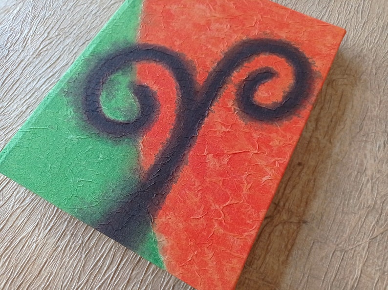 May include: A handmade journal with a green and orange cover. The cover features a black painted design of a ram's horns.