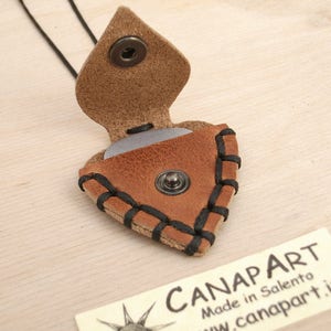 May include: A brown leather guitar pick holder with black stitching and a snap closure. The holder is shaped like a guitar pick and has a silver pick inside. The holder is hanging from a black cord.