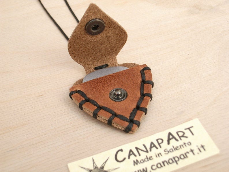 May include: A brown leather guitar pick holder with black stitching and a snap closure. The holder is shaped like a guitar pick and has a silver pick inside. The holder is hanging from a black cord.