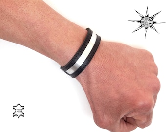 May include: A black leather bracelet with a silver metal strip. The bracelet is on a person's wrist.
