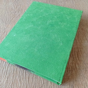 May include: A green hardcover book with a textured cover. The book is lying on a wooden surface.