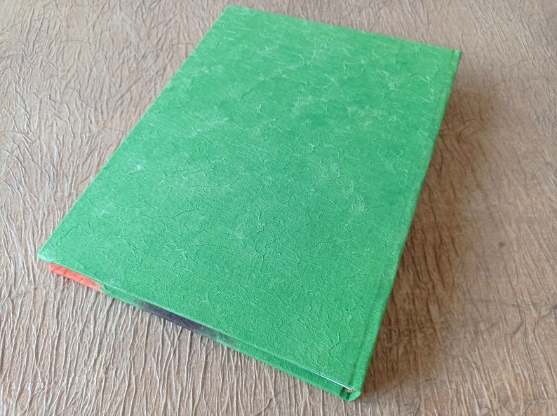 May include: A green hardcover book with a textured cover. The book is lying on a wooden surface.