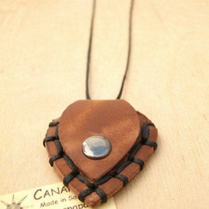 May include: A brown leather heart-shaped pendant with black stitching and a silver metal button. The pendant is attached to a black cord necklace.