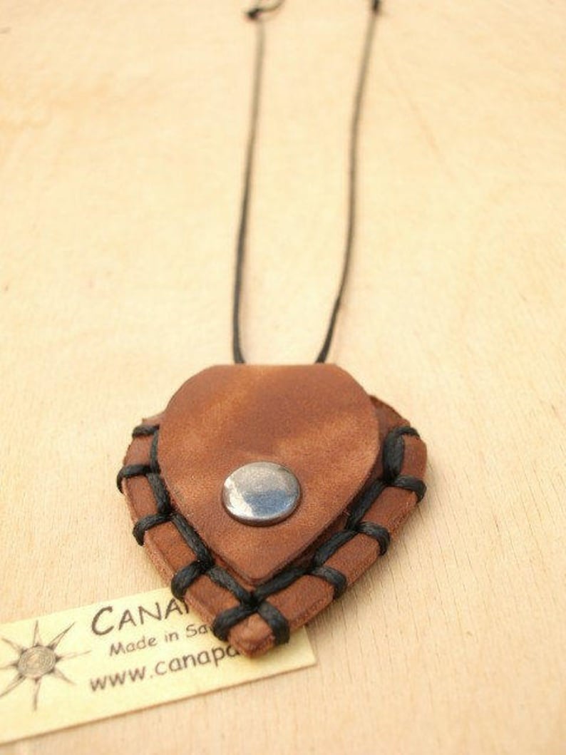 May include: A brown leather heart-shaped pendant with black stitching and a silver metal button. The pendant is attached to a black cord necklace.