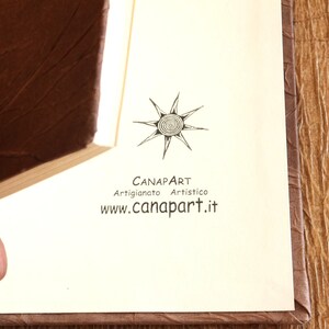 May include: A close-up of a book with a brown leather cover. The book is open to a page with a black and white logo that says "Canapart Artigianato Artistico www.canapart.it"
