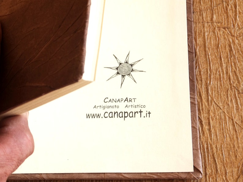 May include: A close-up of a book with a brown leather cover. The book is open to a page with a black and white logo that says "Canapart Artigianato Artistico www.canapart.it"