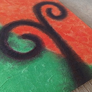 May include: A square canvas with a textured surface. The canvas is painted in two colors, green and orange, with a black abstract design in the center. The design resembles a stylized plant with two swirls.