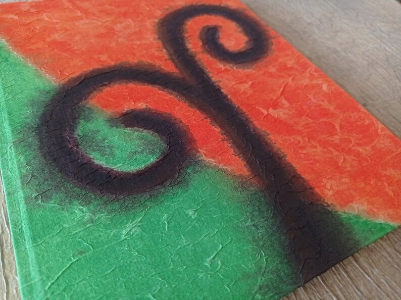 May include: A square canvas with a textured surface. The canvas is painted in two colors, green and orange, with a black abstract design in the center. The design resembles a stylized plant with two swirls.