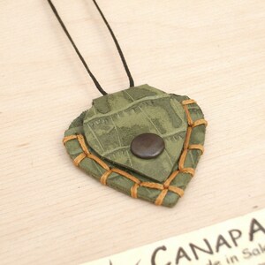 May include: A green leather pendant with brown stitching and a brown button. The pendant is shaped like a heart and is hanging from a black cord.