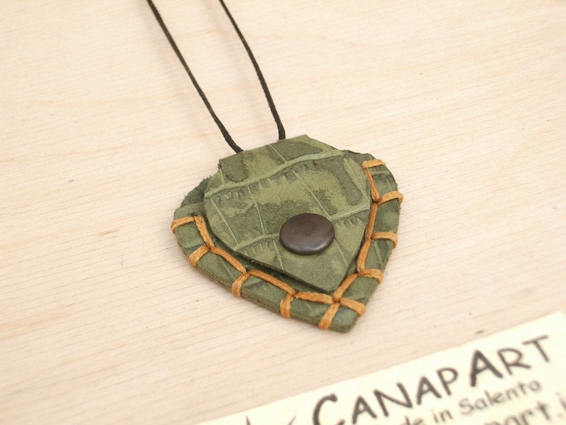 May include: A green leather pendant with brown stitching and a brown button. The pendant is shaped like a heart and is hanging from a black cord.