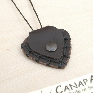 May include: A brown leather guitar pick holder with black stitching and a silver metal snap closure. The holder is attached to a black cord.