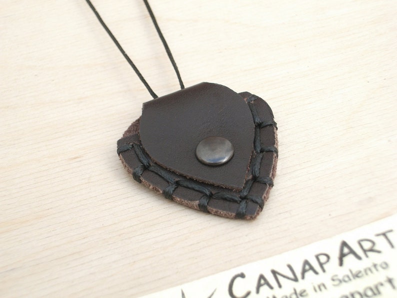 May include: A brown leather guitar pick holder with black stitching and a silver metal snap closure. The holder is attached to a black cord.