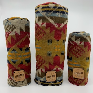May include: Three golf club head covers with a Southwestern geometric pattern in red, gold, blue, and tan. Each cover has a leather tag with the text "REINLAND Golf Co." and a logo. The covers are different sizes.