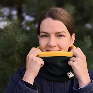 Op de afbeelding: Een vrouw draagt een donkerblauw jack met een groene en gele fleece nekwarmer. De nekwarmer heeft een klein logo aan de zijkant.