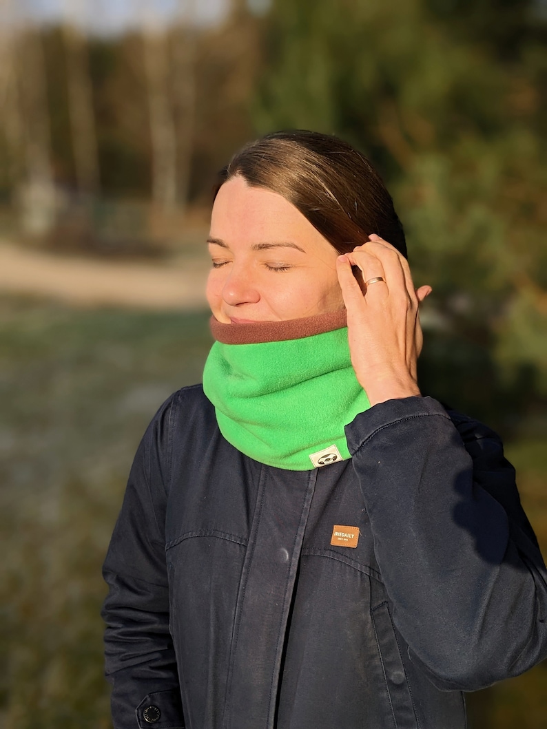 Puede incluir: Un calentador de cuello de forro polar verde y marr&oacute;n. El calentador de cuello es de color verde brillante con un forro interior marr&oacute;n. La persona de la imagen lleva una chaqueta azul oscuro.