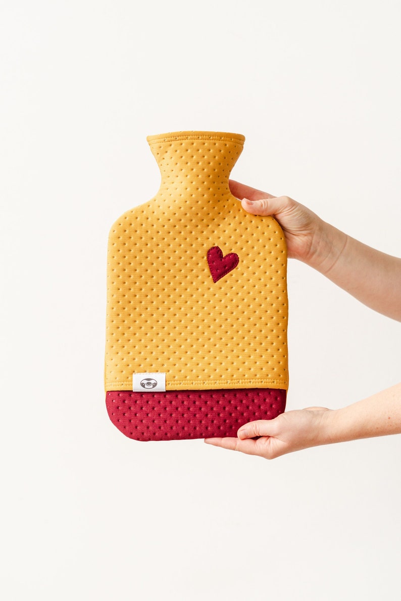May include: A yellow and burgundy hot water bottle with a heart-shaped patch. The bottle has a textured surface and is held by a hand.