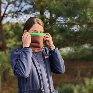 Puede incluir: Un braga de cuello marr&oacute;n y verde es usado. La braga de cuello est&aacute; hecha de un material suave y cubre el cuello y la parte inferior de la cara. La persona lleva una chaqueta azul oscuro con bolsillos y cierres de madera. El fondo es un bosque borroso.