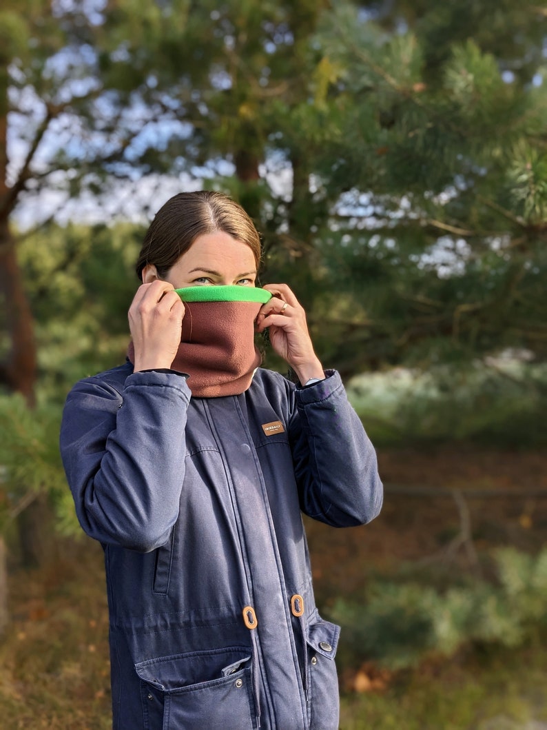 Puede incluir: Un braga de cuello marr&oacute;n y verde es usado. La braga de cuello est&aacute; hecha de un material suave y cubre el cuello y la parte inferior de la cara. La persona lleva una chaqueta azul oscuro con bolsillos y cierres de madera. El fondo es un bosque borroso.