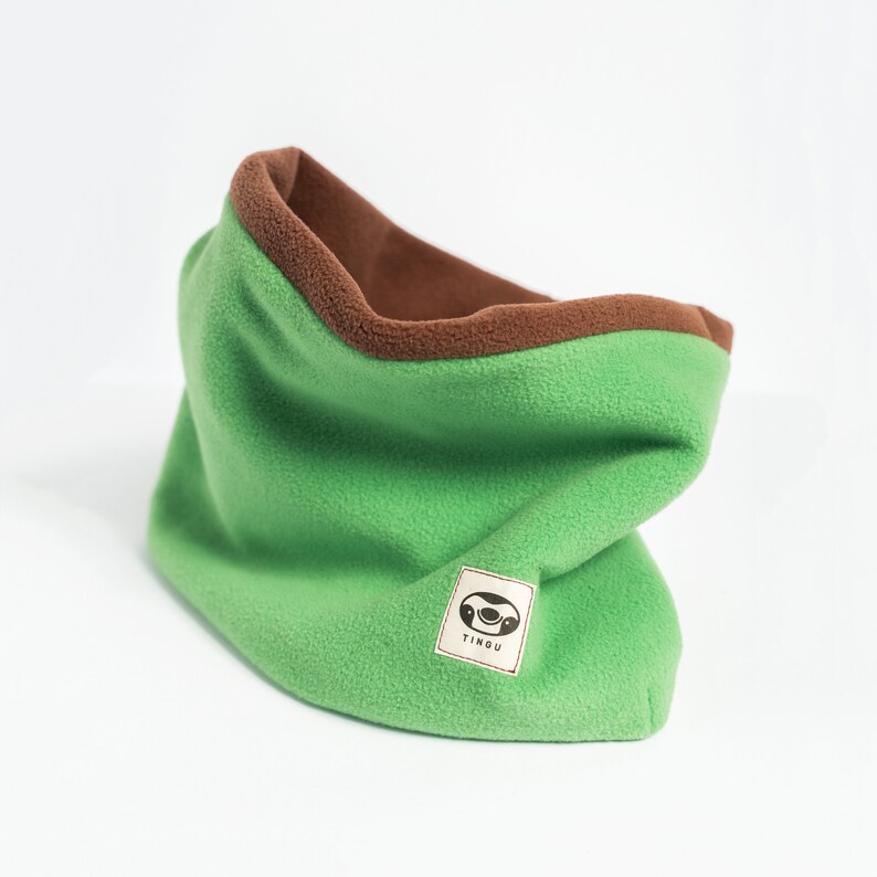 Puede incluir: Un cuello de forro polar verde con interior y ribete marr&oacute;n. El cuello tiene una textura suave y una peque&ntilde;a etiqueta rectangular con un logotipo y la palabra "TINGU".