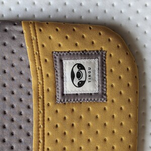 Puede incluir: Primer plano de una esterilla de yoga de color amarillo mostaza con una costura gris y una etiqueta gris que dice "TINGU" con un s&iacute;mbolo yin-yang dentro de un c&iacute;rculo.