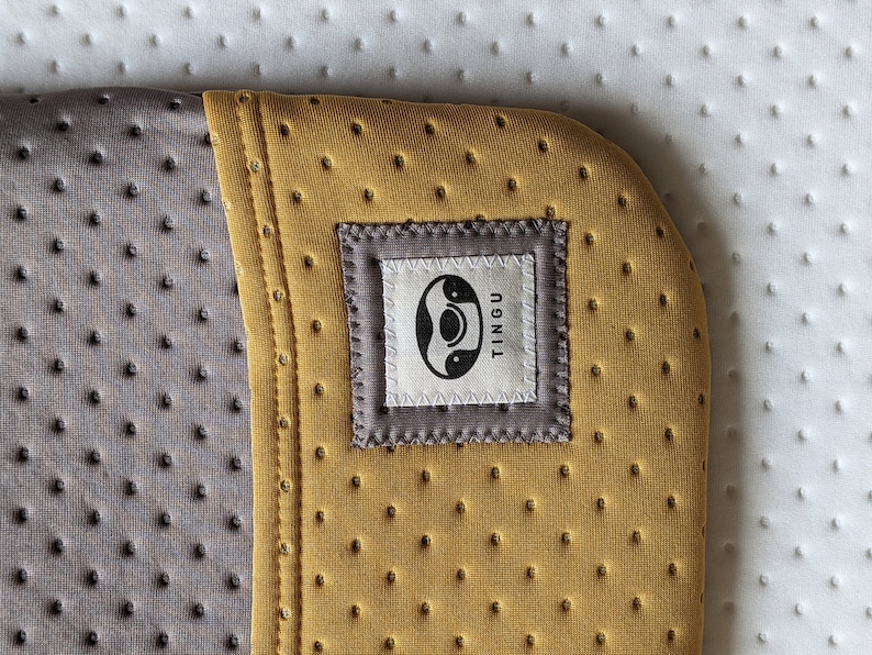 Puede incluir: Primer plano de una esterilla de yoga de color amarillo mostaza con una costura gris y una etiqueta gris que dice "TINGU" con un s&iacute;mbolo yin-yang dentro de un c&iacute;rculo.