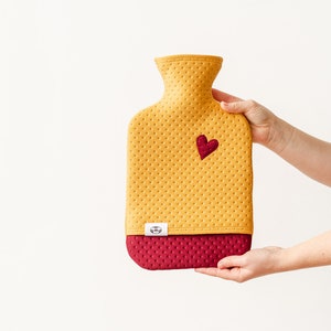 May include: A yellow and burgundy hot water bottle with a heart-shaped patch. The bottle has a textured surface and a small logo on the bottom.