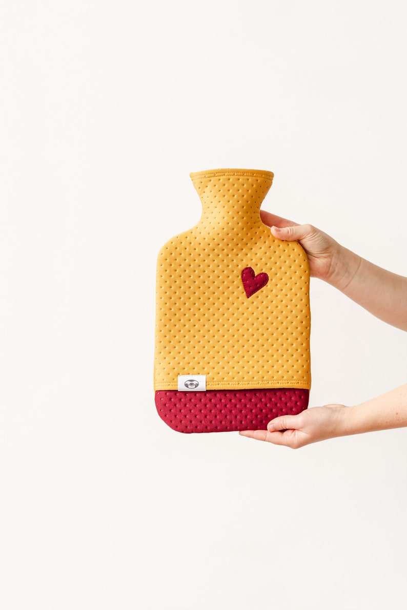 May include: A yellow and burgundy hot water bottle with a heart-shaped patch. The bottle has a textured surface and a small logo on the bottom.