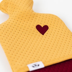 May include: A yellow hot water bottle with a red heart embroidered on the front. The bottle has a red fabric bottom and a white label with a logo.