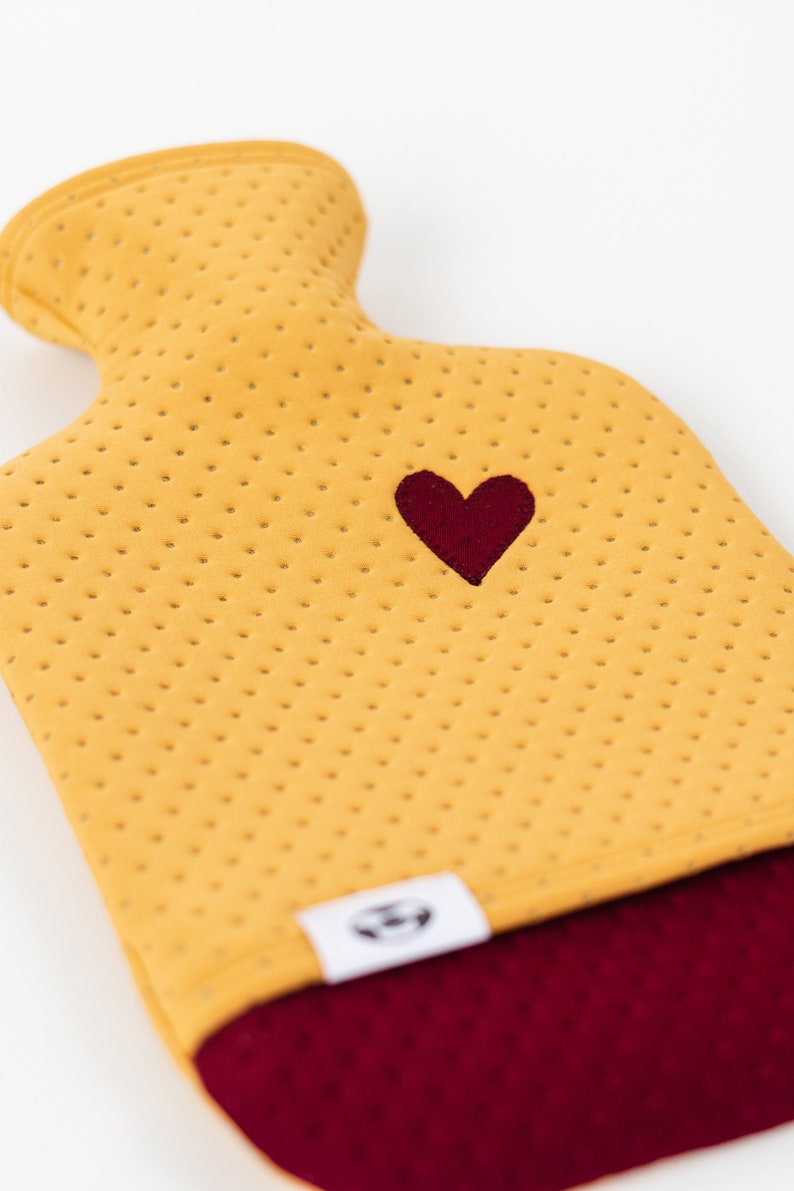 May include: A yellow hot water bottle with a red heart embroidered on the front. The bottle has a red fabric bottom and a white label with a logo.