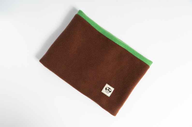 Puede incluir: Un calentador de cuello de forro polar marr&oacute;n con ribete verde. El calentador de cuello tiene una peque&ntilde;a etiqueta cuadrada con un logotipo. Este producto est&aacute; dise&ntilde;ado para mantener el cuello caliente en climas fr&iacute;os. El forro polar es suave y c&oacute;modo.