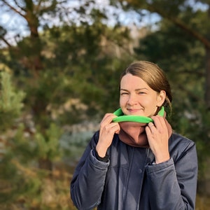 Puede incluir: Un cuello multifuncional multicolor con un dise&ntilde;o verde, marr&oacute;n y verde claro. La persona lleva una chaqueta azul marino. El fondo es una escena de bosque borrosa.