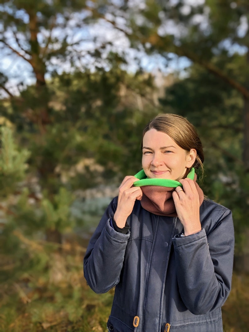 Puede incluir: Un cuello multifuncional multicolor con un dise&ntilde;o verde, marr&oacute;n y verde claro. La persona lleva una chaqueta azul marino. El fondo es una escena de bosque borrosa.
