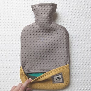 Puede incluir: Una botella de agua caliente gris y amarilla con una cubierta de tela texturizada. La botella tiene una peque&ntilde;a etiqueta con la palabra "YINYU" en ella.