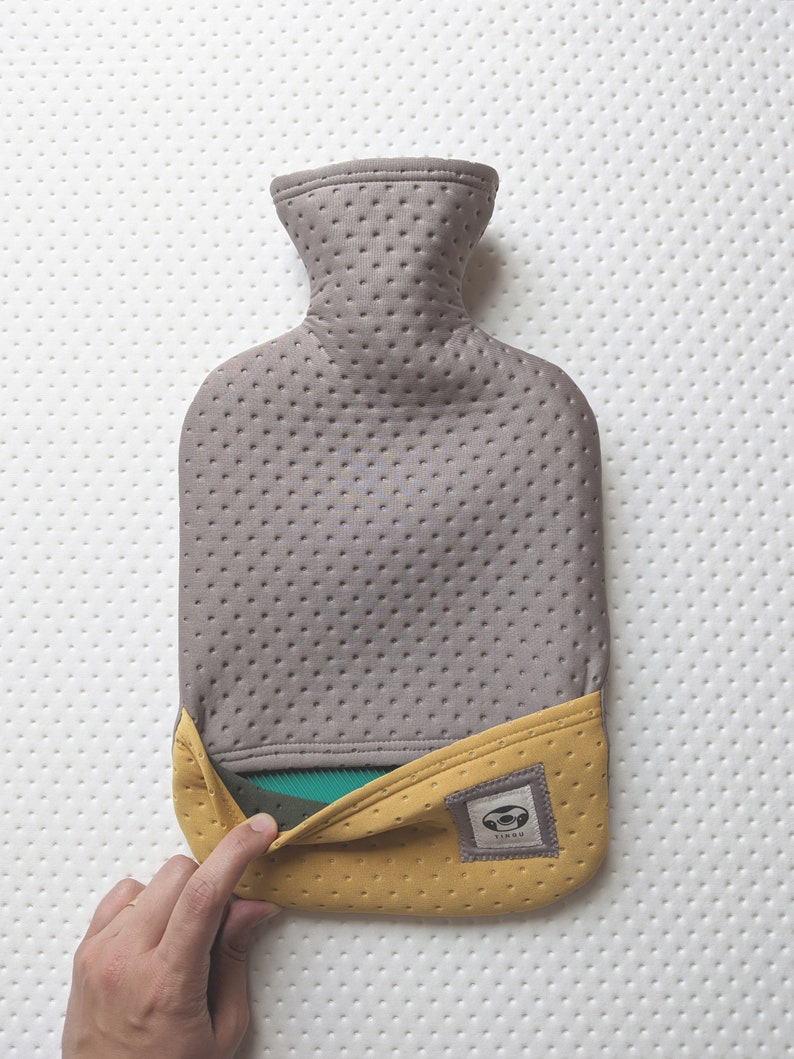 Puede incluir: Una botella de agua caliente gris y amarilla con una cubierta de tela texturizada. La botella tiene una peque&ntilde;a etiqueta con la palabra "YINYU" en ella.