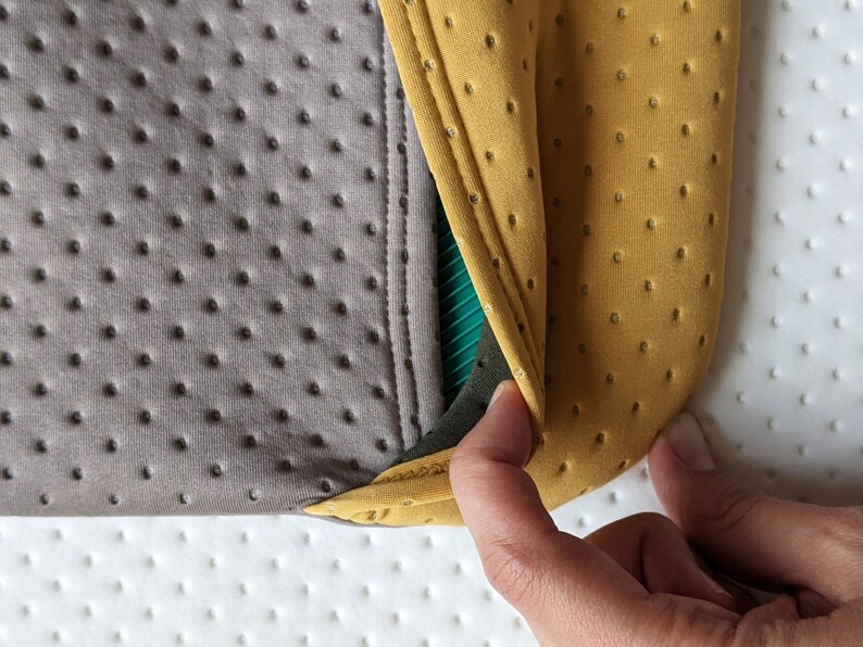 Puede incluir: Primer plano de una tela gris y amarilla con un patr&oacute;n texturizado. La tela est&aacute; doblada y muestra una capa verde y negra debajo.