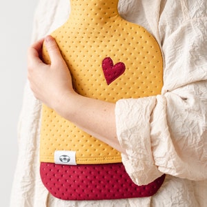 May include: A yellow and burgundy quilted hot water bottle with a heart-shaped patch. The bottle has a textured surface and a small label with a sloth design.