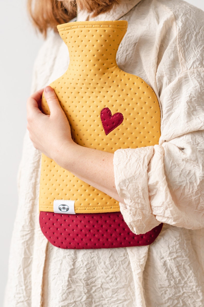 May include: A yellow and burgundy quilted hot water bottle with a heart-shaped patch. The bottle has a textured surface and a small label with a sloth design.