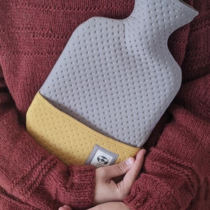 Puede incluir: Una botella de agua caliente gris y amarilla con una superficie texturizada y un logotipo de perezoso. La botella est&aacute; siendo sostenida por una persona que lleva un su&eacute;ter marr&oacute;n.