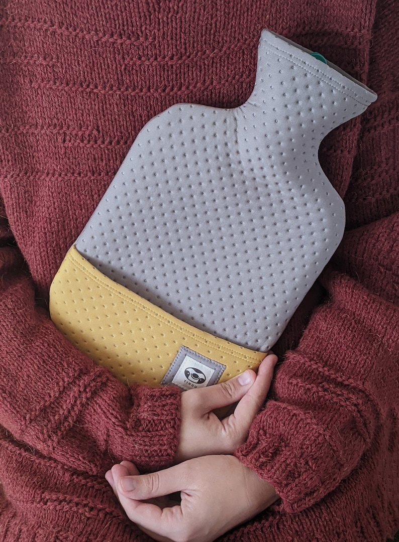 Puede incluir: Una botella de agua caliente gris y amarilla con una superficie texturizada y un logotipo de perezoso. La botella est&aacute; siendo sostenida por una persona que lleva un su&eacute;ter marr&oacute;n.