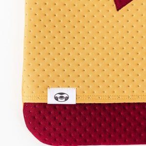 May include: A yellow fabric with a red heart and a white label with a sloth face. The fabric has a textured pattern of small dots. The bottom of the fabric is a darker red color with a similar textured pattern.